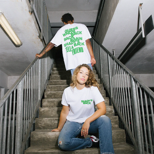 Two models on a staircase, one seated wearing the 'Shady's Back' T-shirt with the front slogan, while the other stands showing the back print, celebrating the 20th anniversary of The Eminem Show.