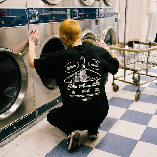 Person in laundromat inserting laundry into a washing machine while wearing 'Shady Cleaners T-shirt' with 'Clean out my closet' text and a hanger graphic on the back.