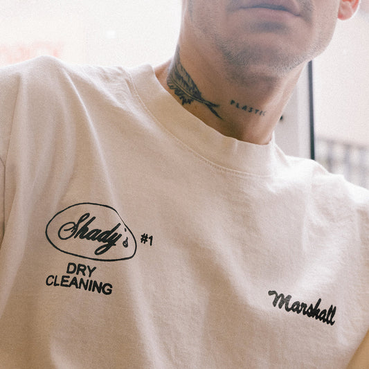 Close-up view of the 'Shady Cleaners T-shirt' in cream with 'Shady’s #1 Dry Cleaning Marshall' chest print, modeled by a person with a neck tattoo.