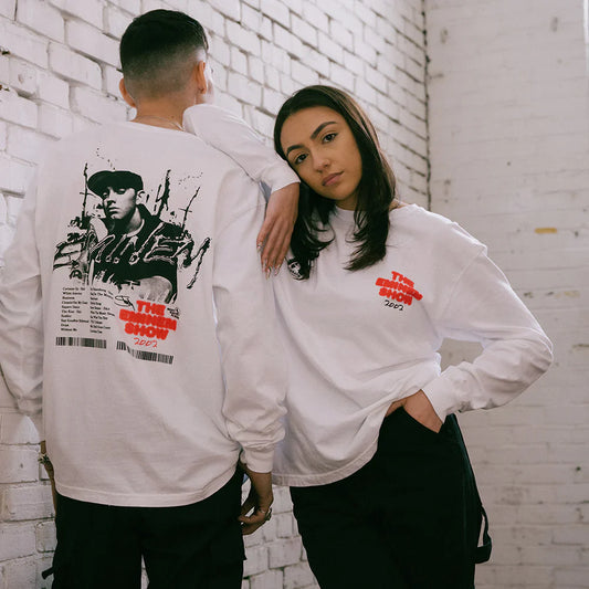 A man and woman posing back-to-back in a white brick-walled setting, sporting 'The Eminem Show 2002' long sleeve shirts featuring iconic album artwork.